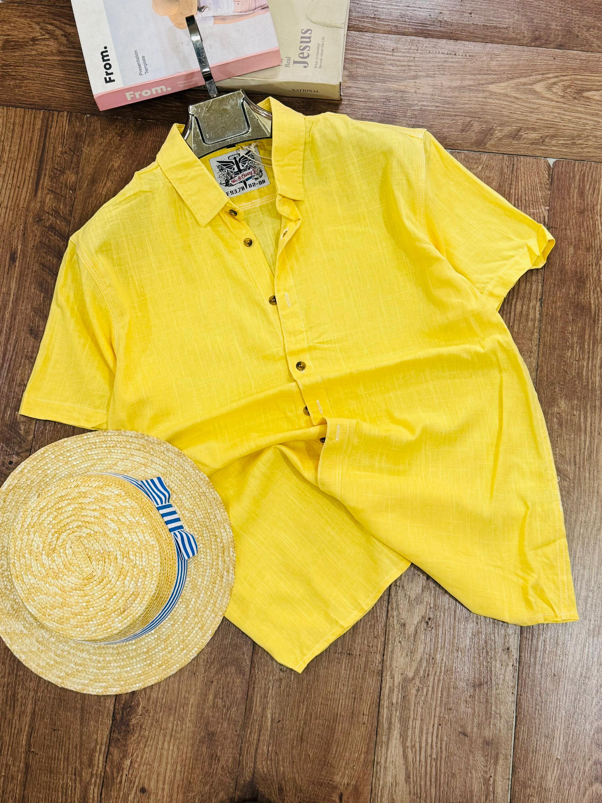 A bright yellow short-sleeve button-up shirt laid flat on a wooden floor, styled with a straw hat and a book.